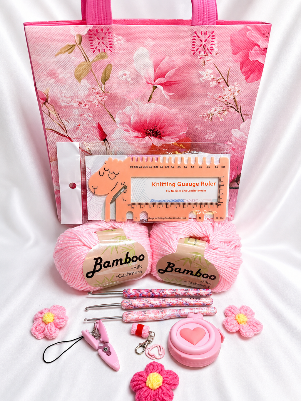 A pink crochet gift arranged on a white satin background, including a large pink tote bag, two balls of pink yarn labeled Bamboo, a pattern booklet, a ruler, three floral crochet hooks, a pink stitch marker, a small pink heart-shaped tool, and several pink flower-shaped accessories.